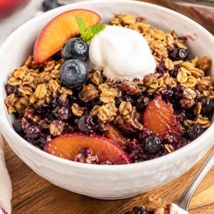 blueberry peach crisp in a white bowl with a scoop of vanilla nice cream.