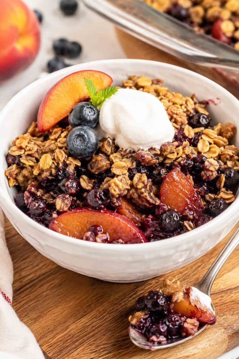 blueberry peach crisp in a white bowl with a scoop of vanilla nice cream.