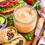 spicy vegan mayonnaise in a small jar with a spoon and a sandwich and wraps on table beside.