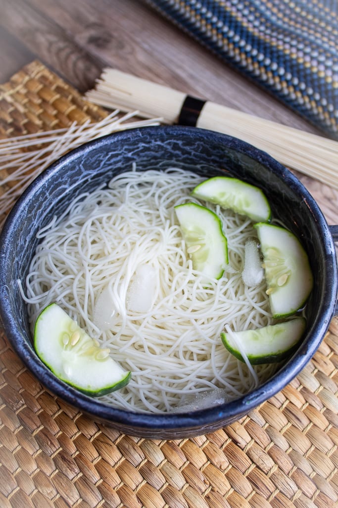 blue bowl of cooked somen noodles in ice water with cucumber slices floating