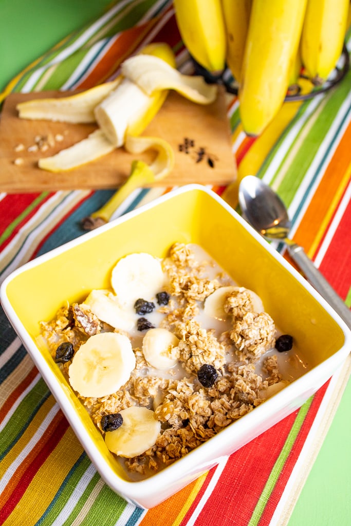 peanut butter granola cereal in bright yellow bowl topped with bananas and blueberries