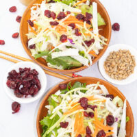 overhead of 2 wooden bowls filled with napa cabbage salad with chopsticks, sunflower seeds, and dried cranberries surrounding on white counter