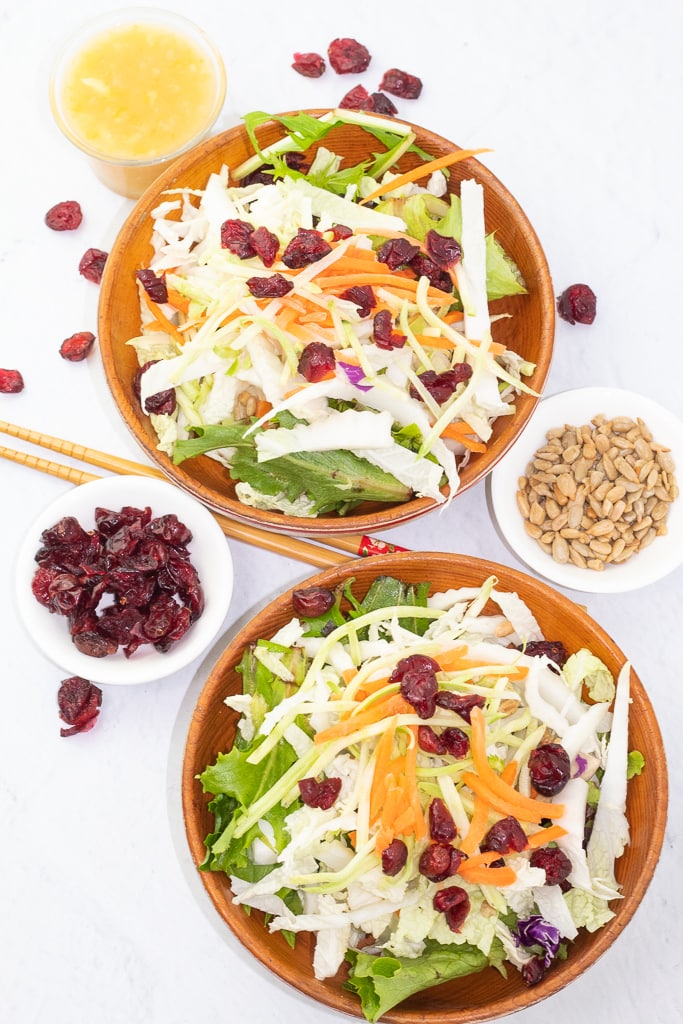 overhead of 2 wooden bowls filled with napa cabbage salad with chopsticks, sunflower seeds, and dried cranberries surrounding on white counter