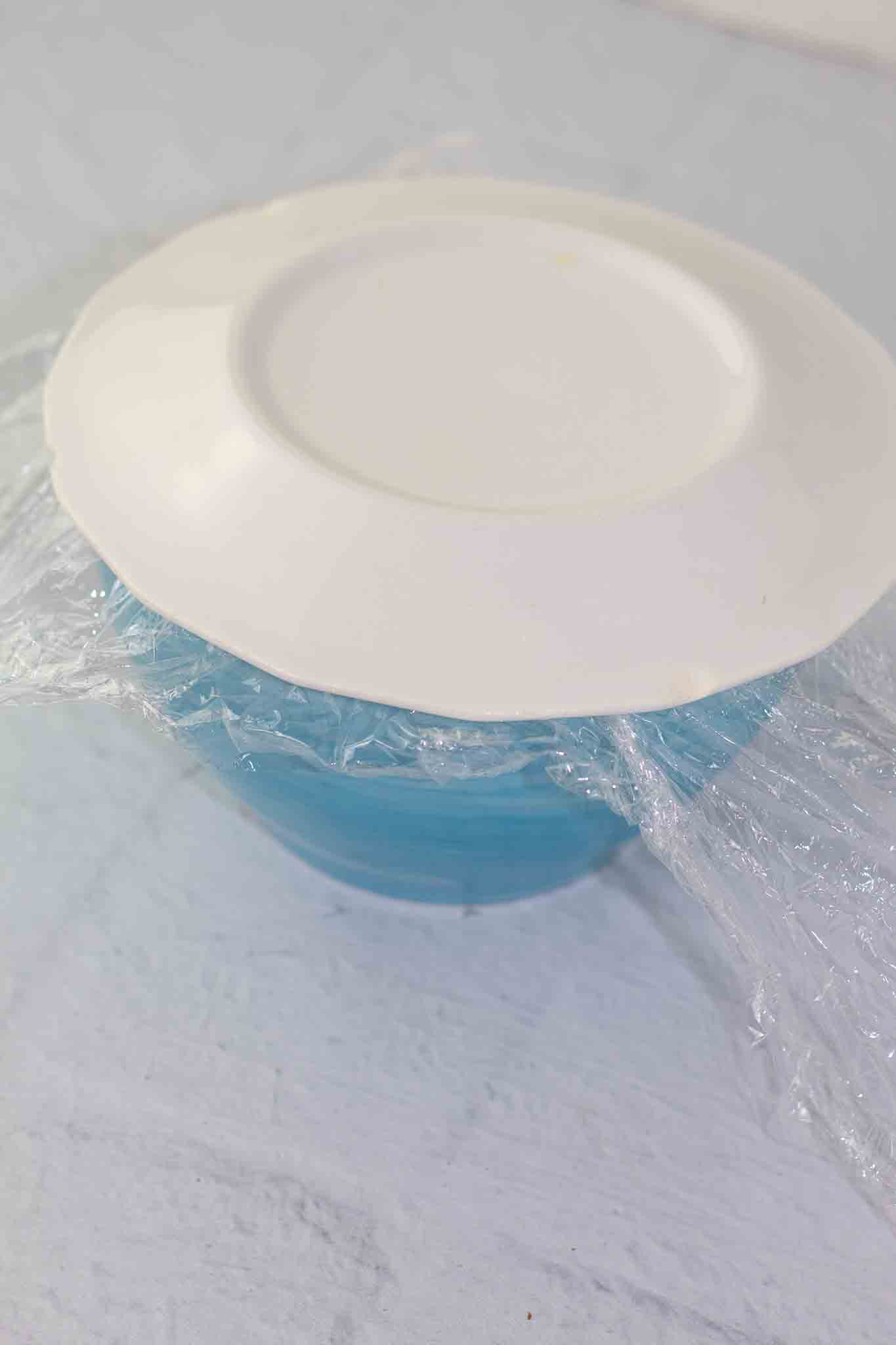 small blue bowl with plastic wrap and cheese ball covered with a white plate to be inverted onto a platter.