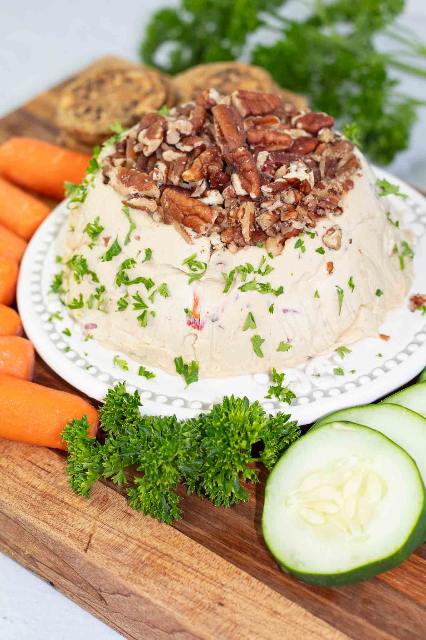 wooden board filled with cucumber slices, baby carrots, and crackers with a vegan cheese ball topped with pecan pieces in the middle.