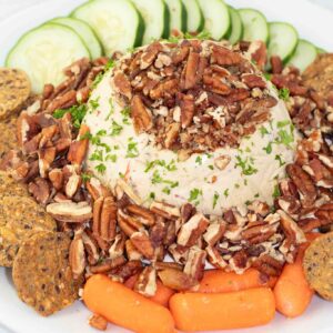 white platter with a vegan cheese ball in the middle that is covered with nuts and surrounded by veggies and crackers.