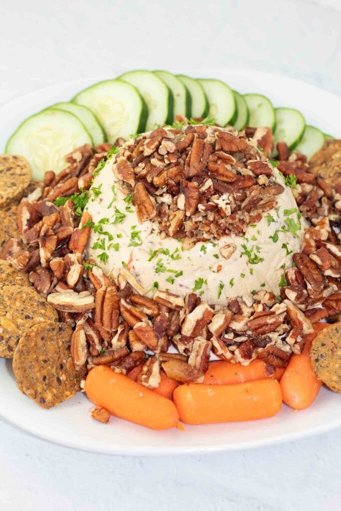 white platter with a vegan cheese ball in the middle that is covered with nuts and surrounded by veggies and crackers.