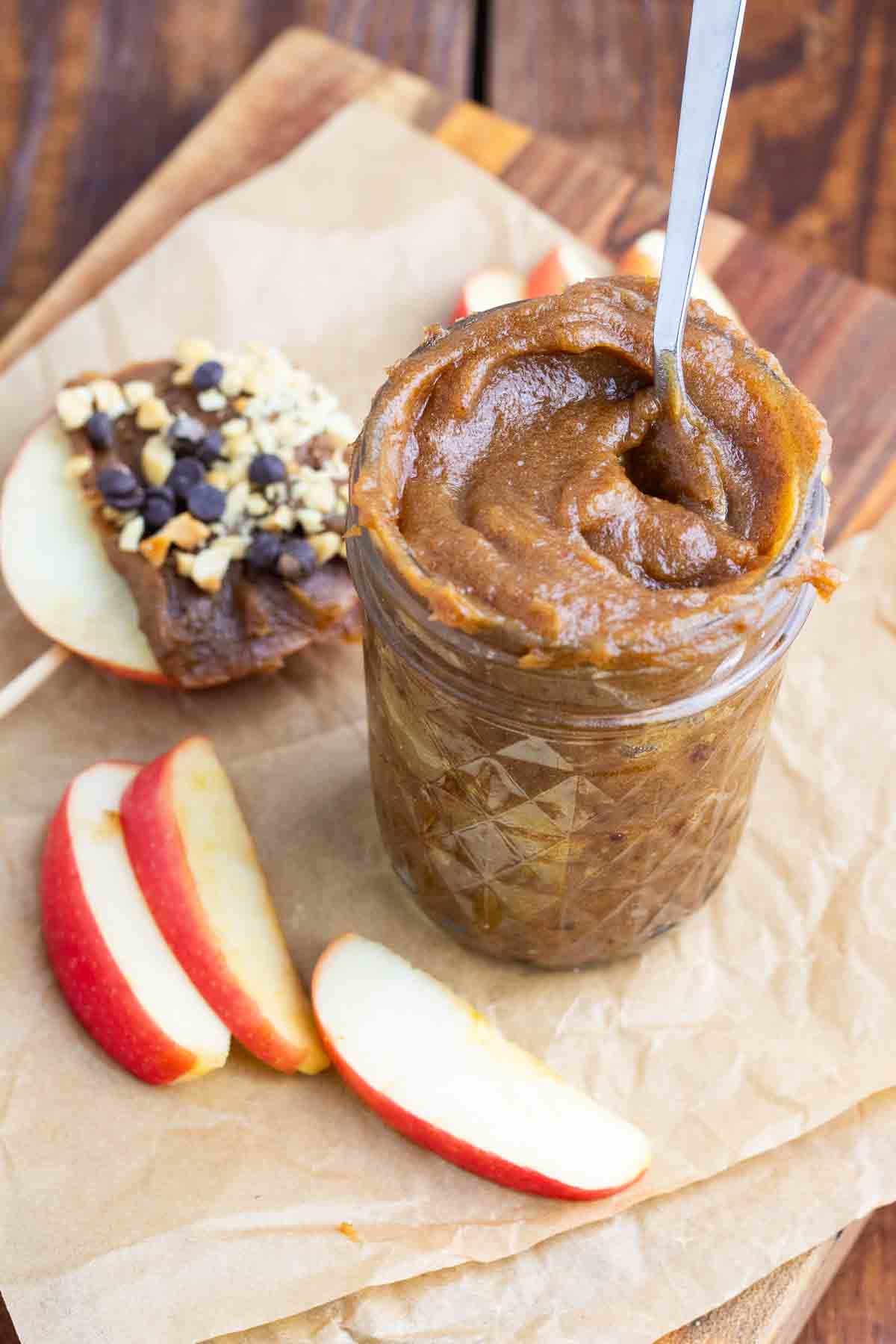 small glass jar filled with vegan caramel made from dates with a spoon inside jar and apple slices below.