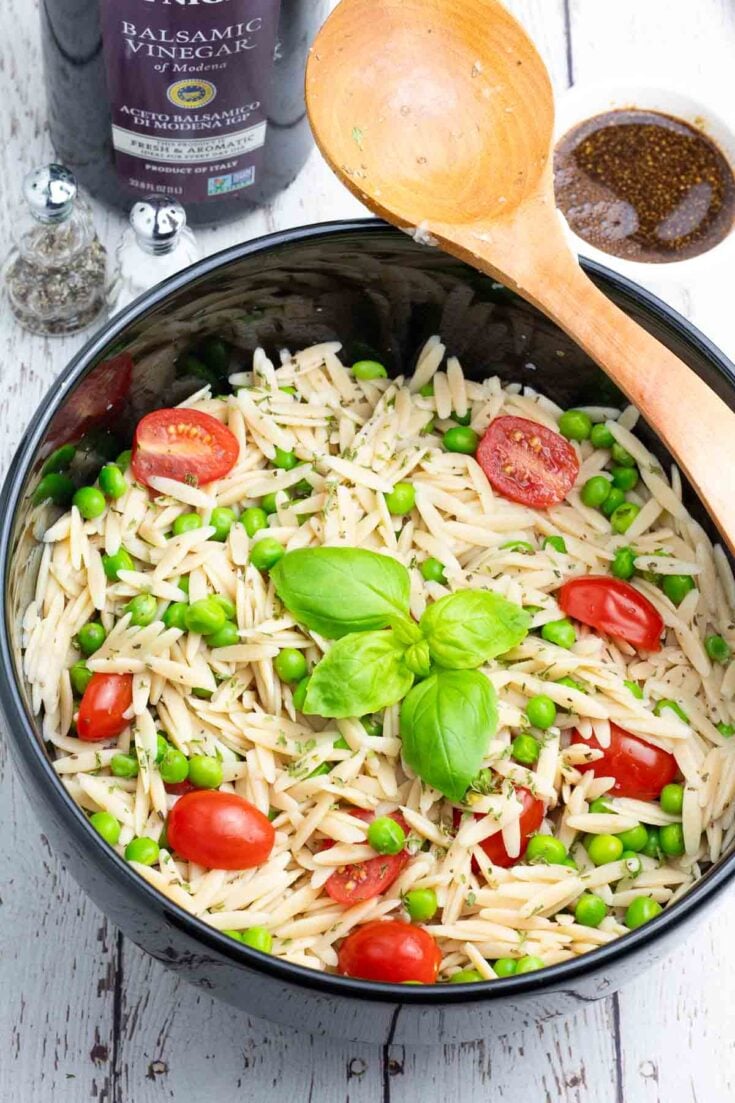 black serving bowl filled with orzo pasta salad topped with tomatoes and basil with a wooden spoon resting on side.