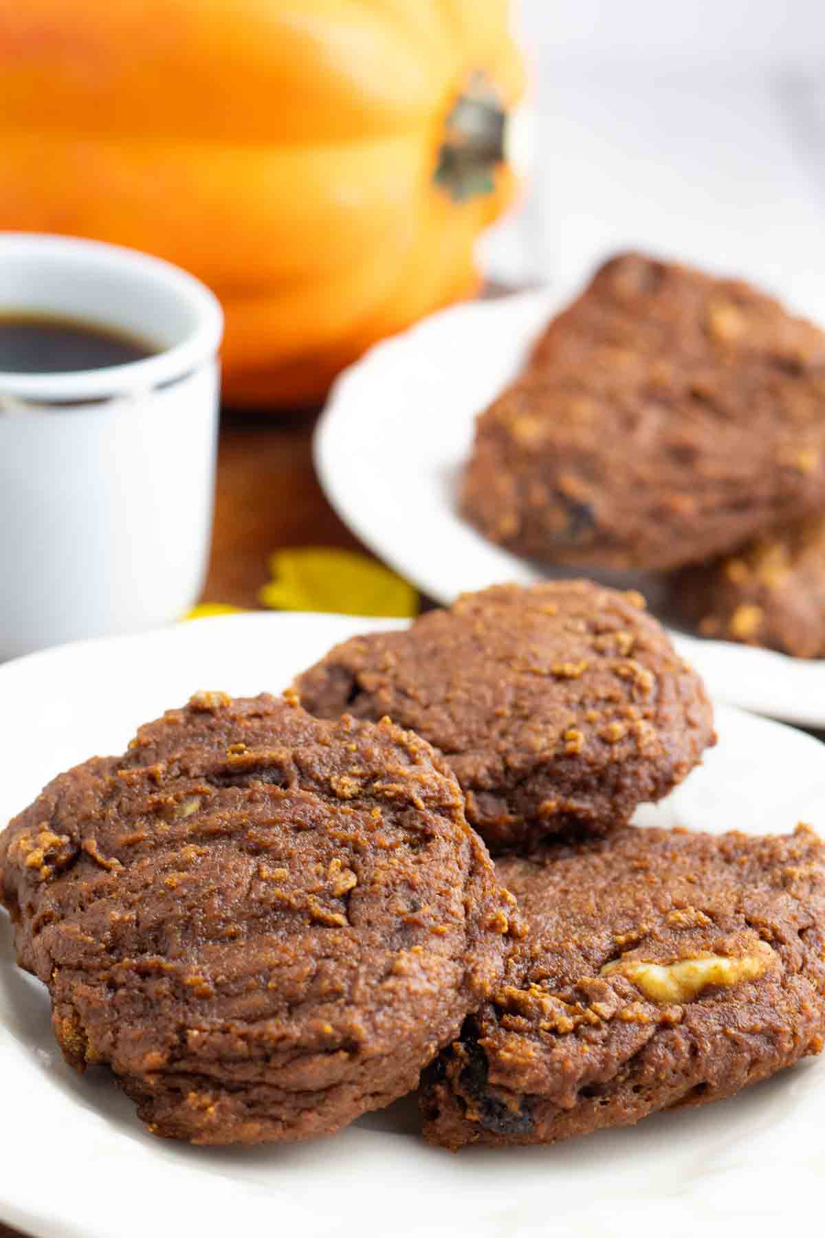 3 vegan pumpkin cookies on a white plate with a cup of coffee and whole pumpkin in background.