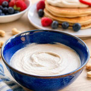 vanilla cashew cream in a blue bowl with pancakes and fruit in background.