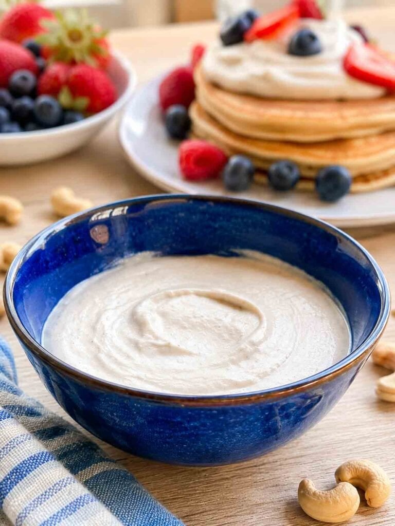 vanilla cashew cream in a blue bowl with pancakes and fruit in background.