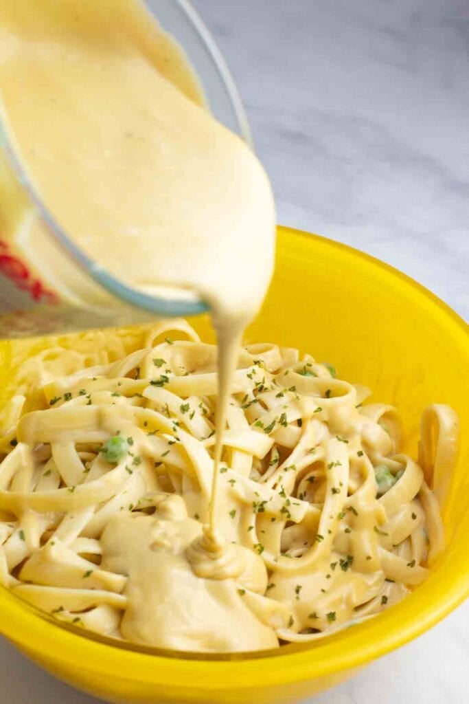cashew vegan alfredo being poured from a glass gravy dish into a bright yellow bowl of pasta.
