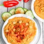 white bowl filled with roasted red pepper hummus with cucumber slices and red peppers in the background.
