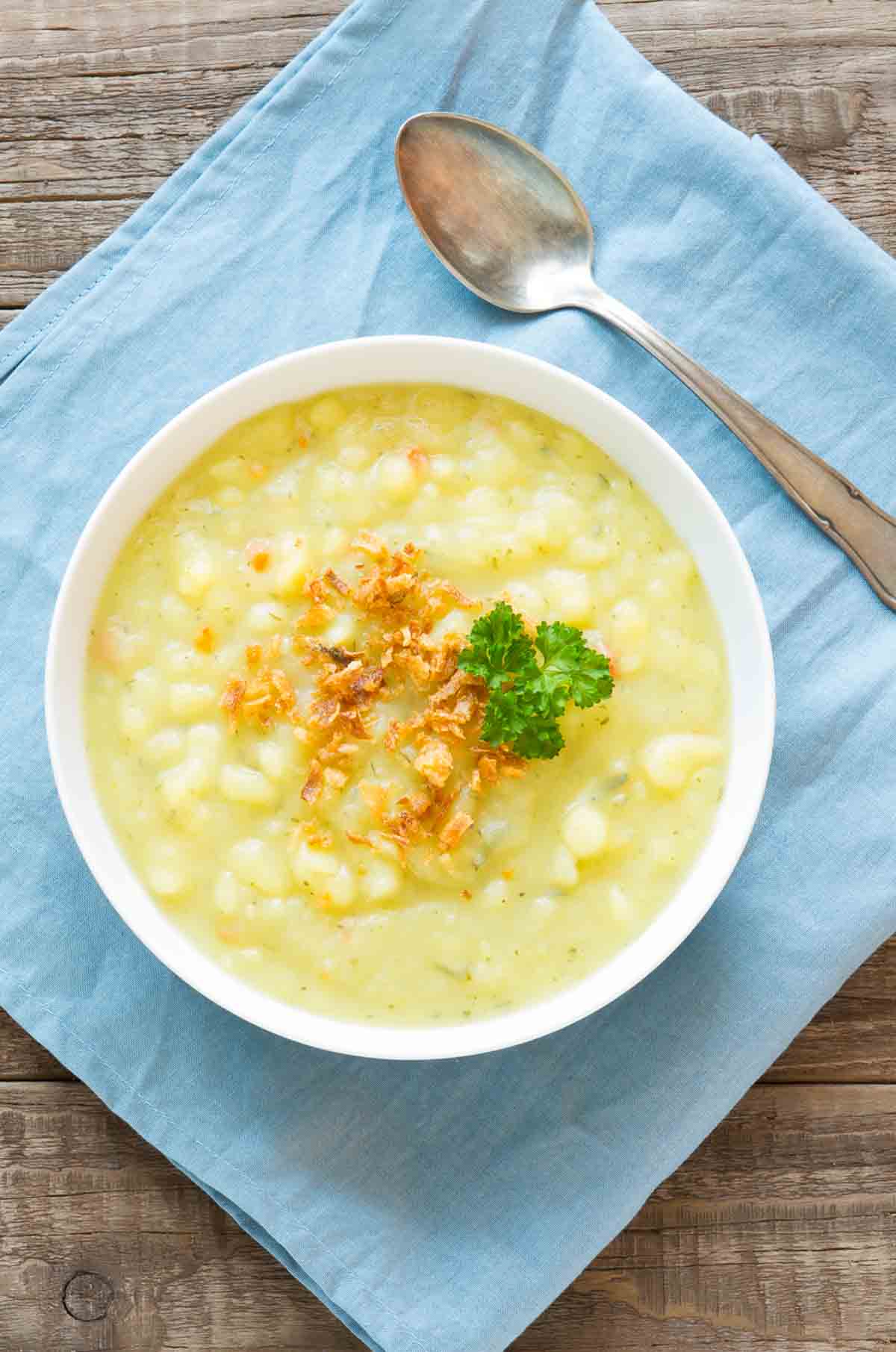 white bowl of homemade potato soup topped with parsley on a blue napkin with a spoon.