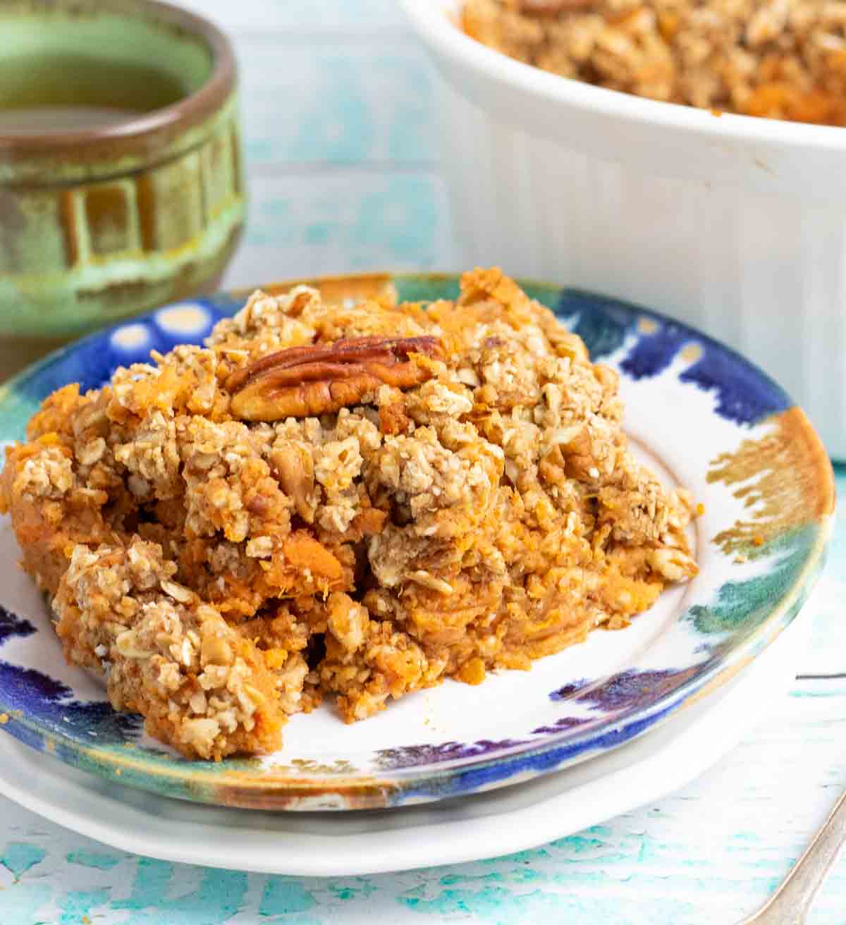 a slice of vegan sweet potato casserole on a colorful plate with a roasted pecan on top and coffee in the background.