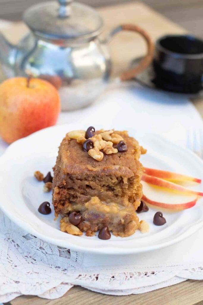 white dessert plate with a slice of vanilla crockpot cake that is topped with crushed walnuts and chocolate chips.