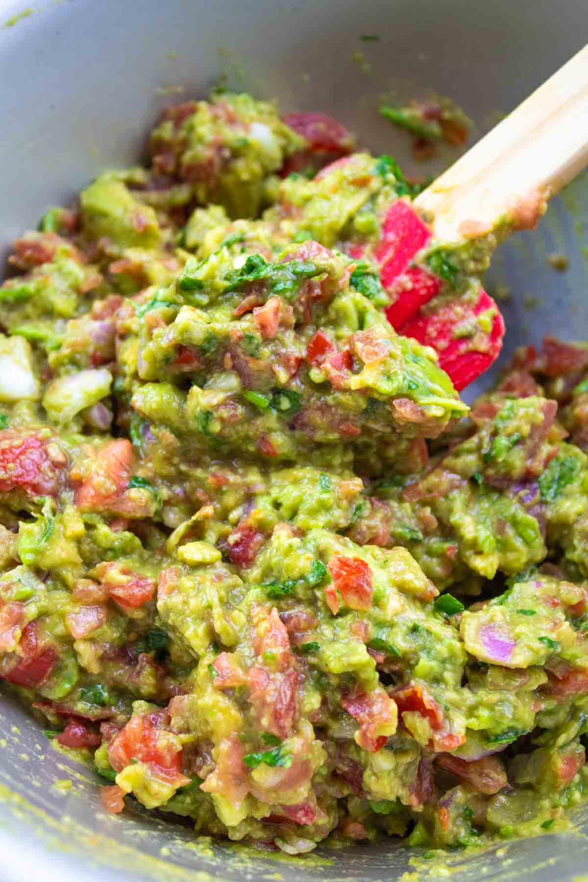 mixing bowl with mashed avocado, tomatoes, onion, cilantro, and cumin being stirred with red spatula.