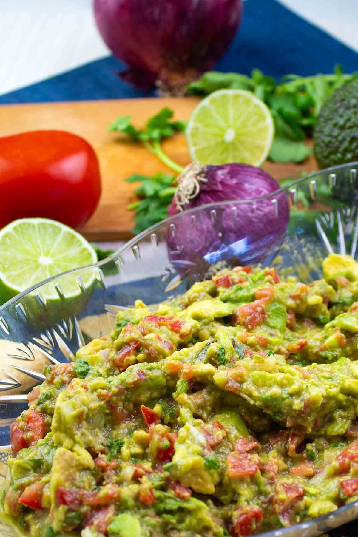 oval clear serving bowl full of avocado guacamole with vegetables in the background.