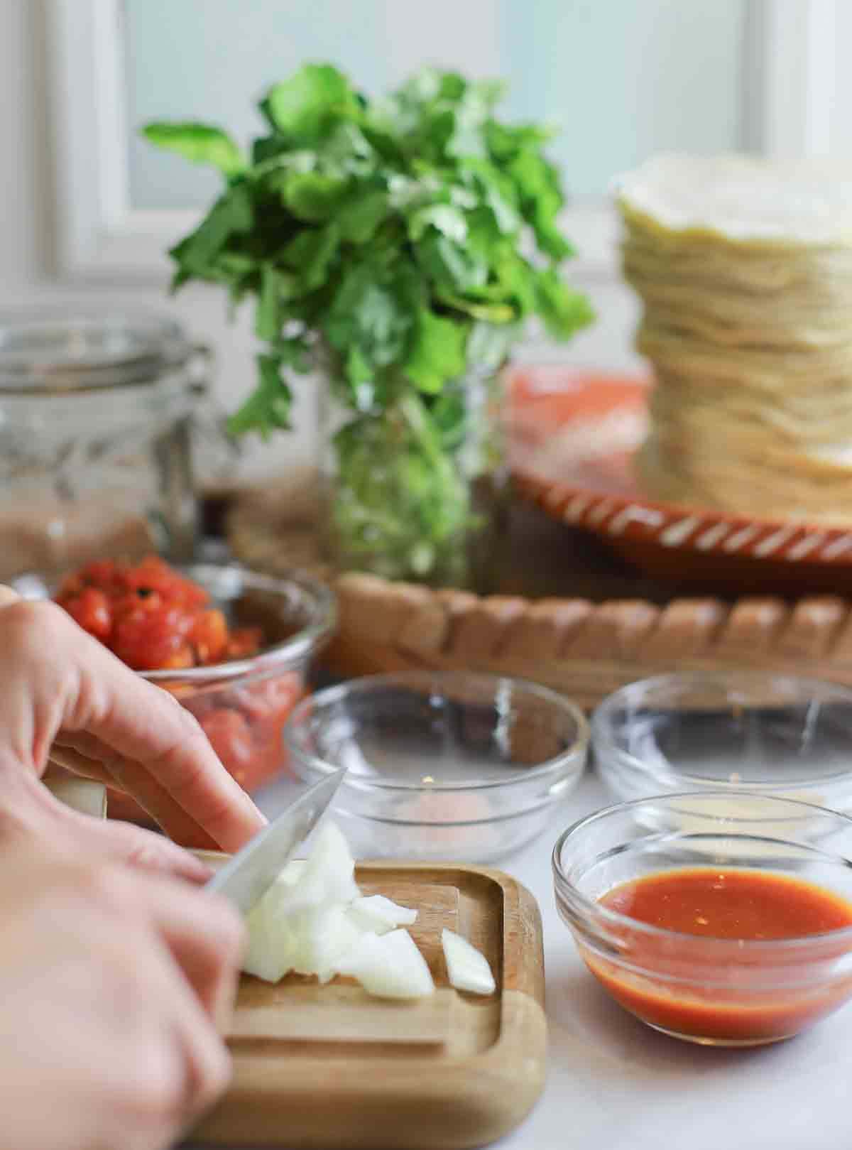 person's hands chopping onion on a cutting board with tomatoes and cilantro in background.