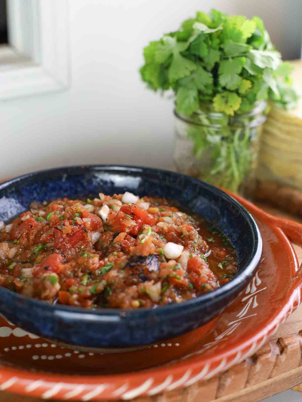 big blue bowl full of homemade red salsa with jar of cilantro in background.