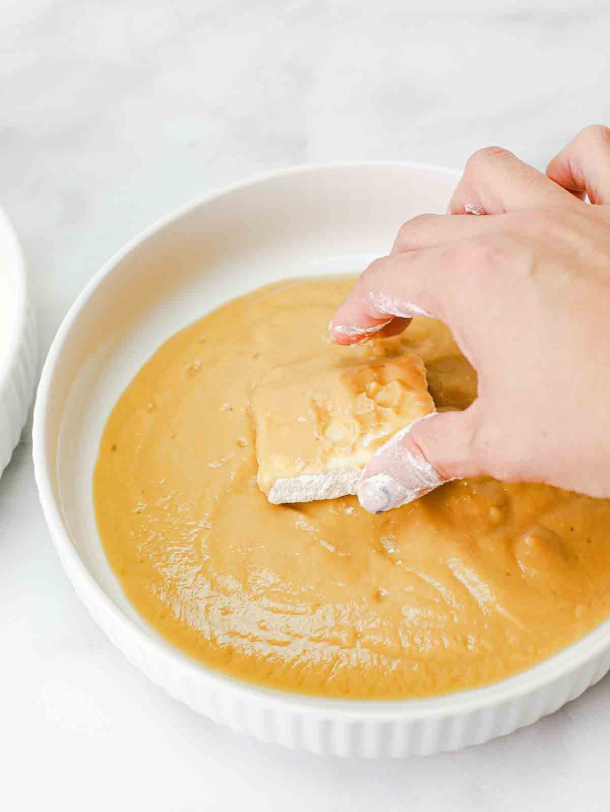 slice of tofu being rolled in a bowl of chickpea batter.