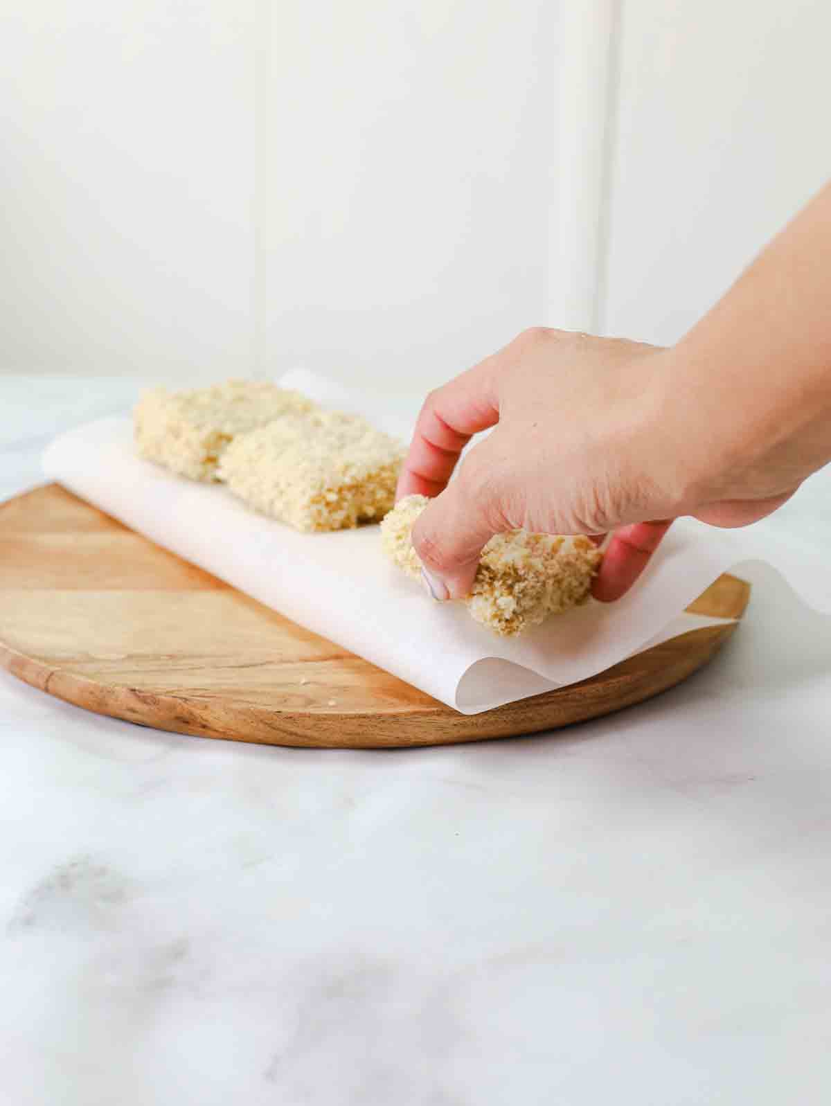persons hand placing breaded tofu on a cutting board with parchment paper.