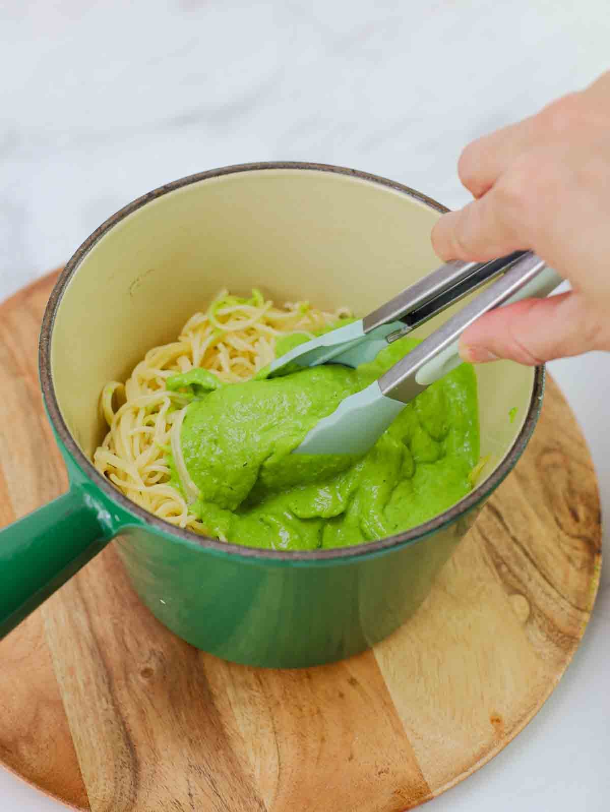 green verde sauce being mixed into cooked spaghetti noodles with tongs.