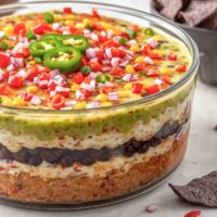 large clear, glass, round dish full of 7-layer dip topped with tomatoes, onions, and jalepenos.