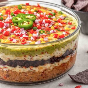 large clear, glass, round dish full of 7-layer dip topped with tomatoes, onions, and jalepenos.