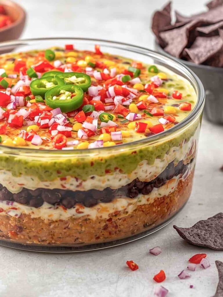 large clear, glass, round dish full of 7-layer dip topped with tomatoes, onions, and jalepenos.