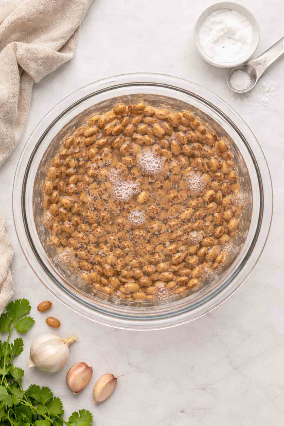 glass bowl filled with dry pinto beans covered with water and baking soda in the background.