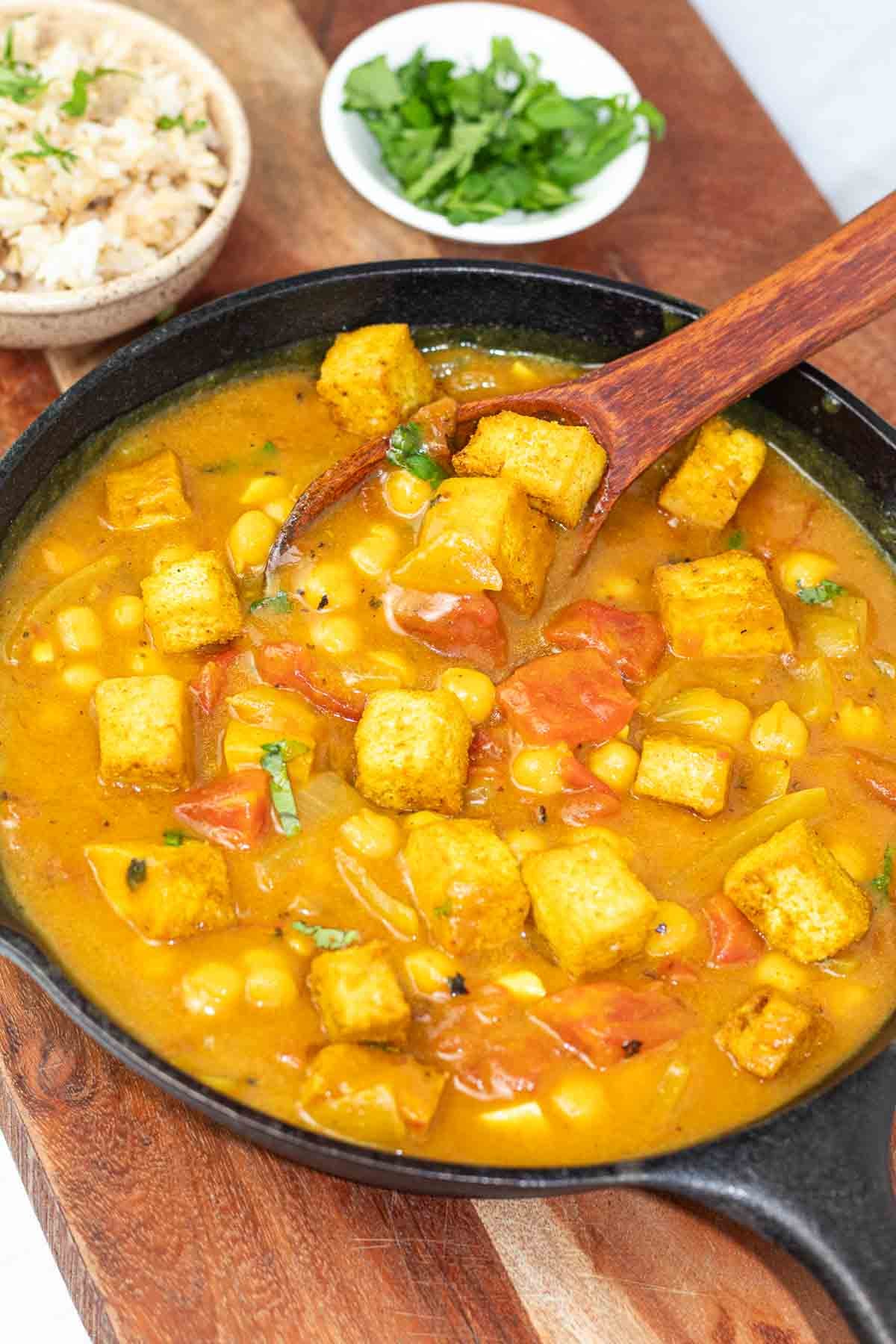 black iron skillet with tofu chickpea curry being served with a wooden spoon.