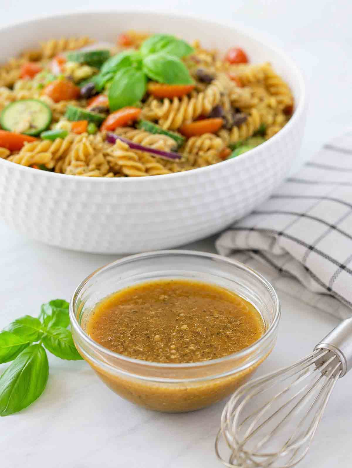 homemade maple dijon dressing in a small glass bowl with a pasta salad behind it showing it can be used to season pasta salad.