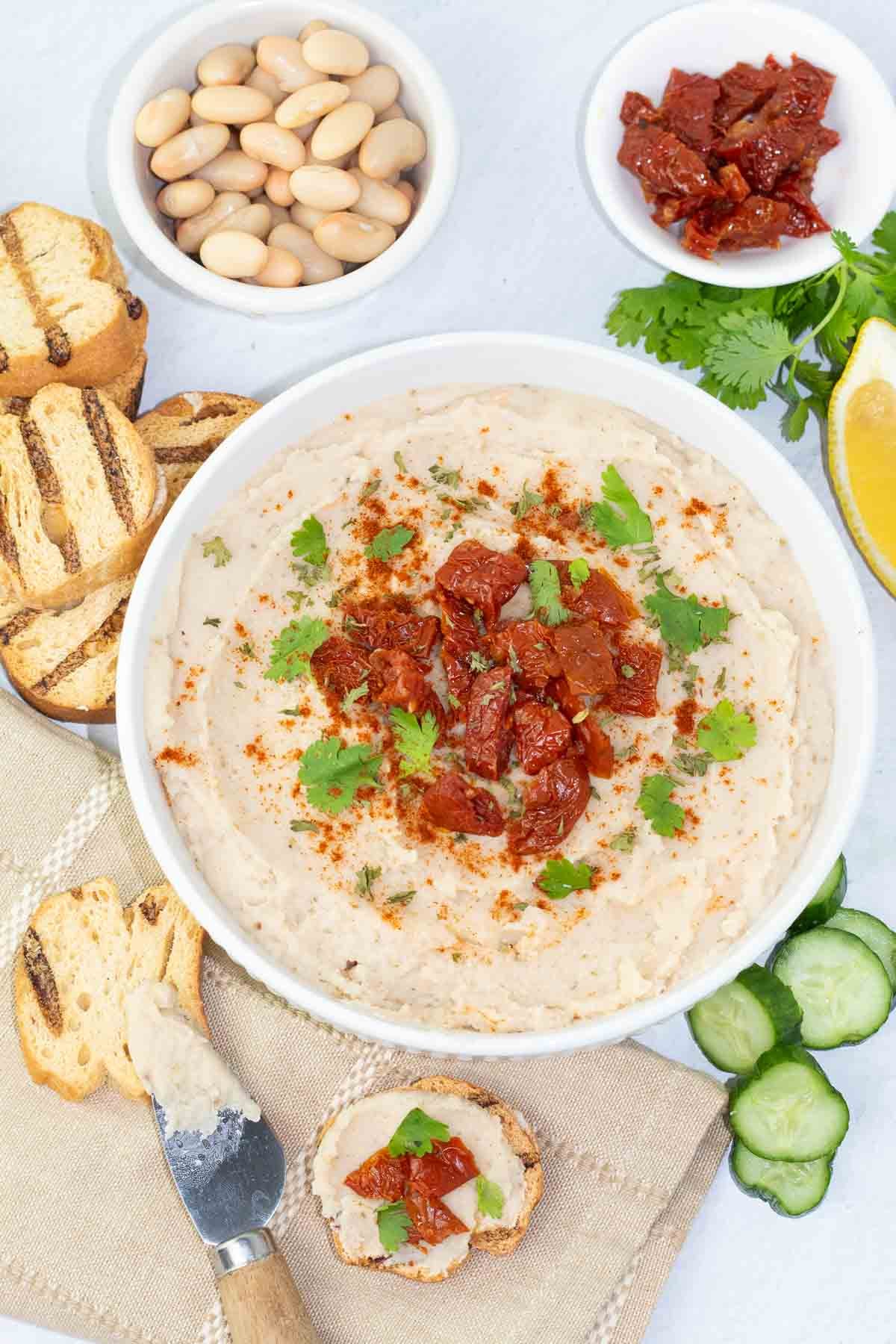 white bean dip in a bowl with some ingredients surrounding the bowl like great northern beans, sundried tomatoes, and crusty bread.