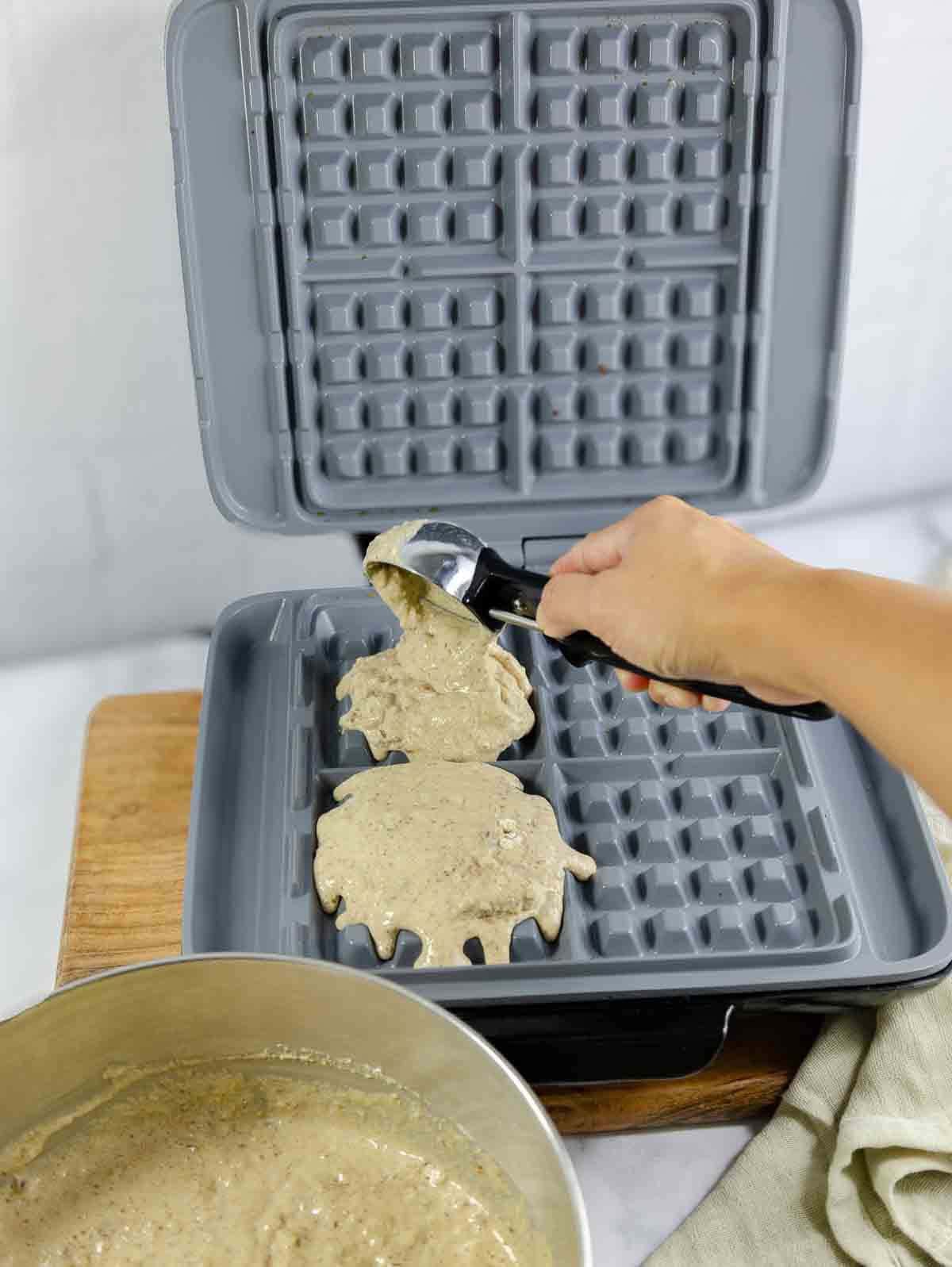 open waffle maker with a person using an icecream scoop for waffle batter.