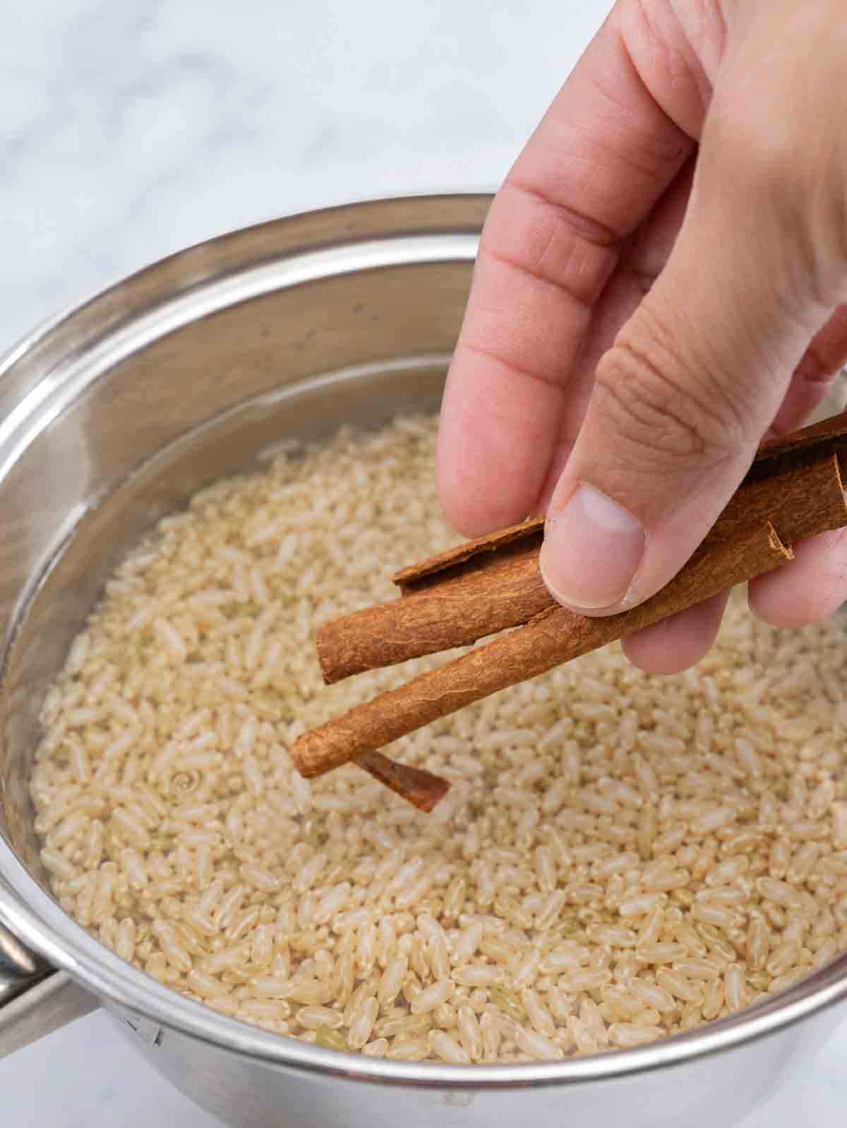 stainless pot with brown rice and an hand placing cinnamon sticks in it.