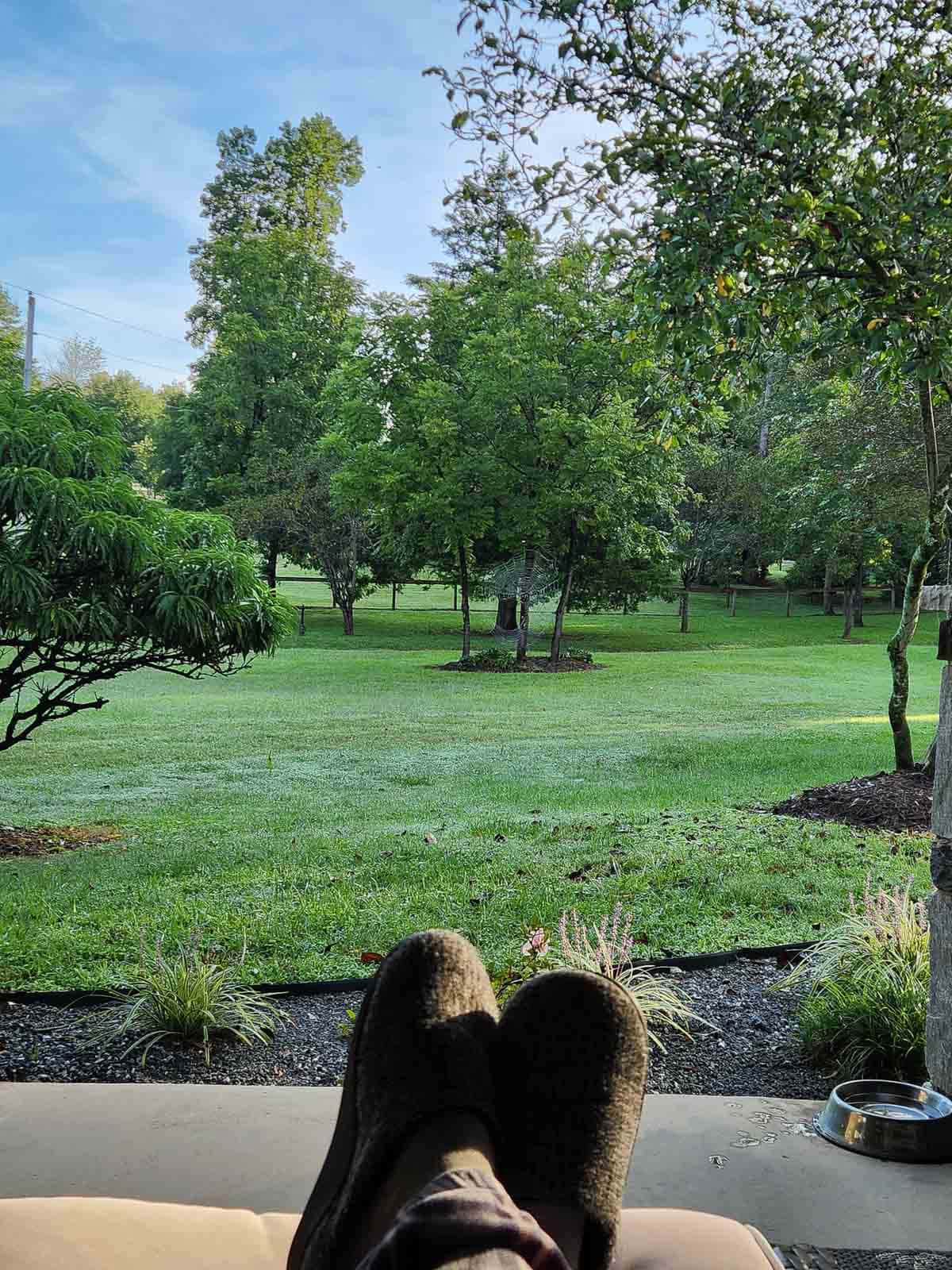 person's feet sitting on porch looking in a green yard.