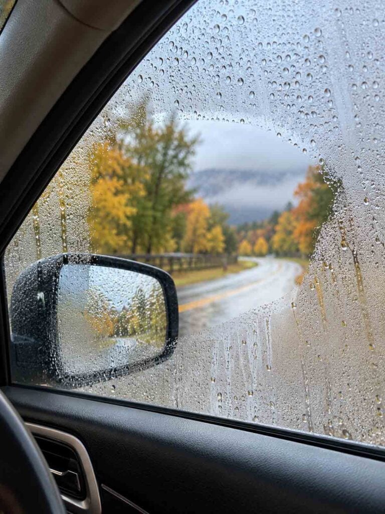 car side window is covered with condensation and raindrops, making the beautiful country road and autumn trees outside look blurry and difficult to see.