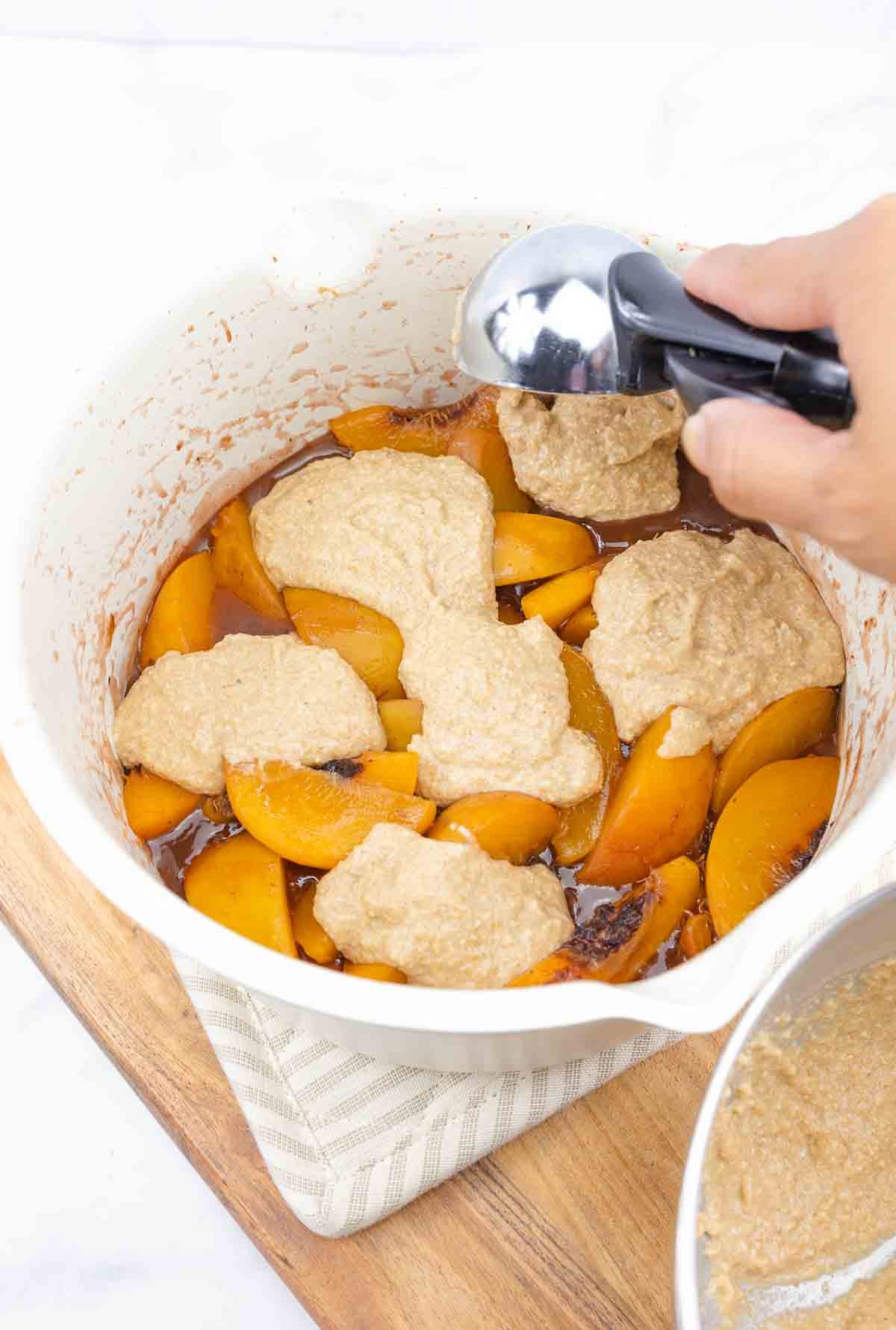 wet batter being dolloped over cooked peaches with an ice cream scoop.