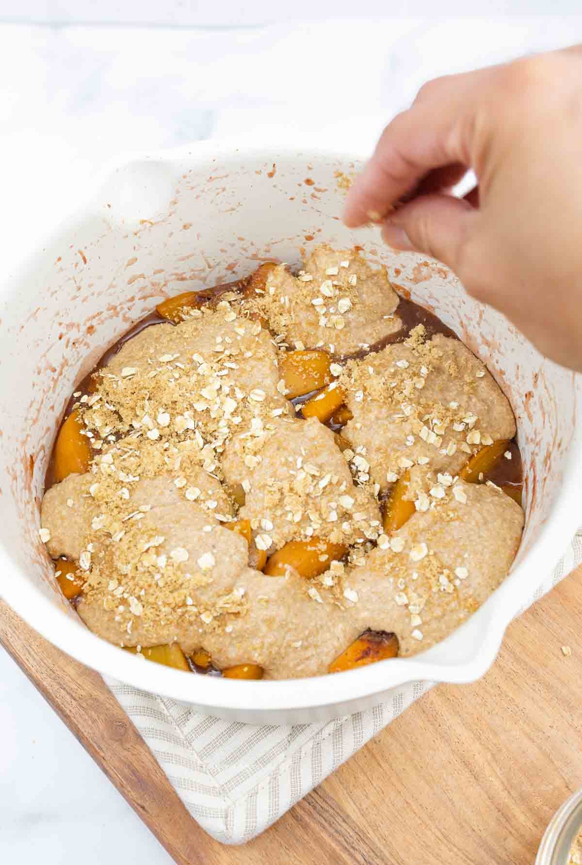 someone sprinkling brown sugar and oats on top of a cobbler before baking.