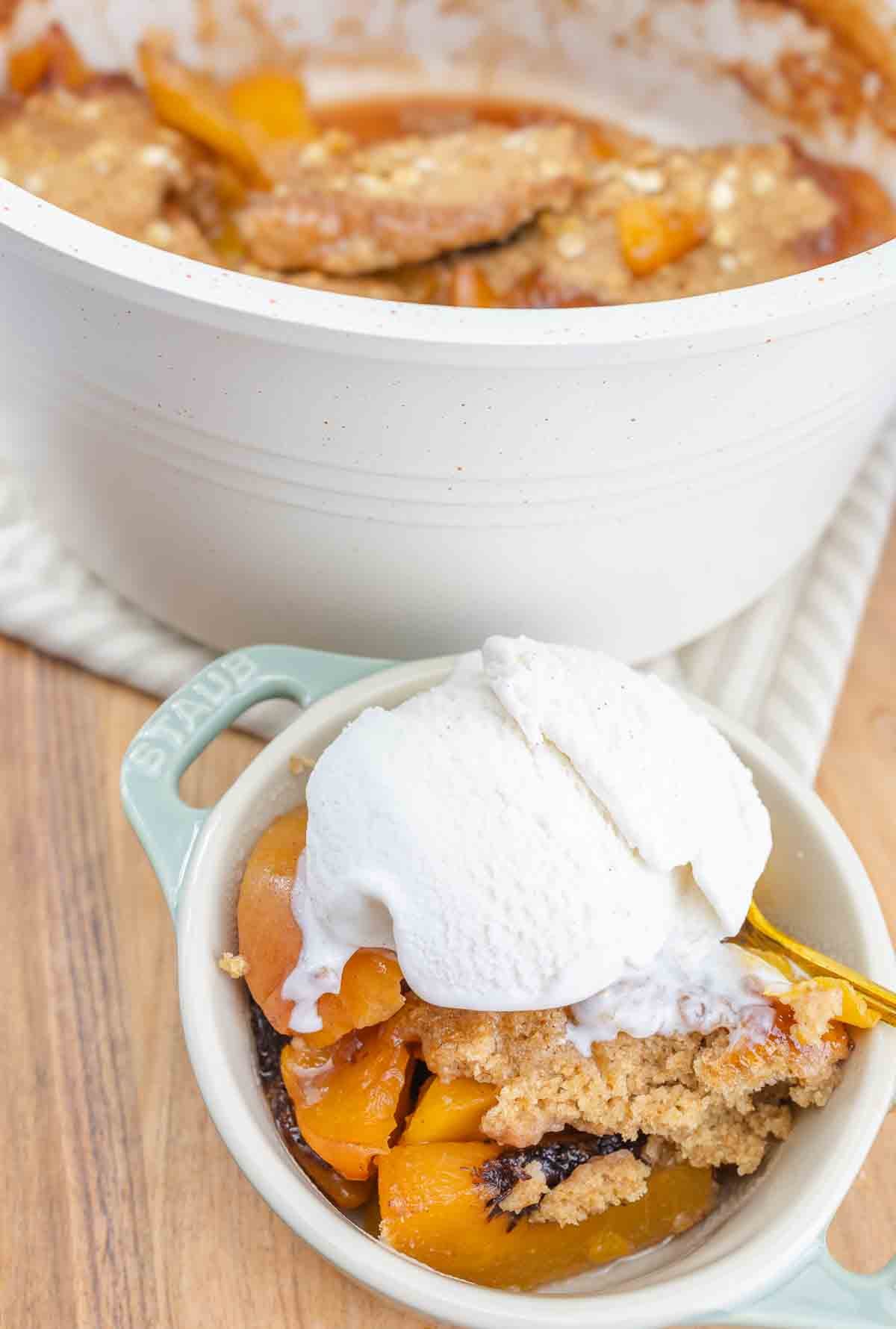 bowl of peach cobbler with a scoop of vanilla ice cream on top and baking pan of cobbler in the background.
